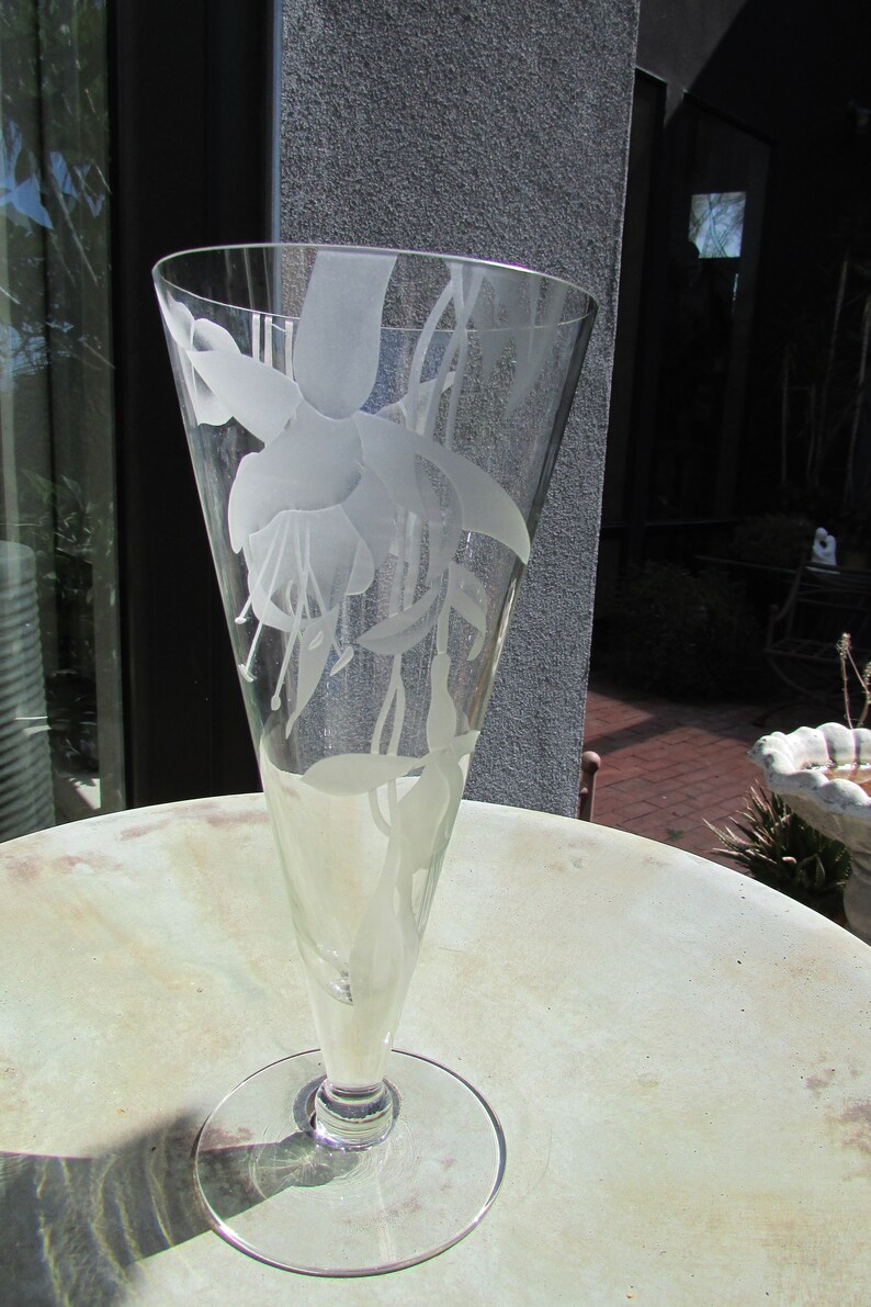 Puede incluir: Un jarr&oacute;n de cristal transparente con la parte superior acampanada y una base redonda. El jarr&oacute;n est&aacute; grabado con un dise&ntilde;o floral con flores y hojas de fucsia. El jarr&oacute;n est&aacute; sobre una mesa redonda.