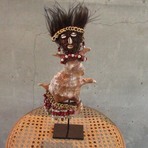 May include: A handmade figurine of a person with a dark brown hair and a shell body. The figurine is decorated with red and white beads, gold chain, and seashells. The figurine is standing on a black stand.