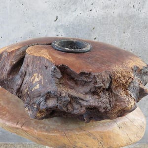 May include: A rustic, handcrafted wooden bowl with a dark, circular insert. The bowl is made from a single piece of wood, showcasing natural grain and texture. The wood has a rich, warm brown color, with a rough, organic edge.