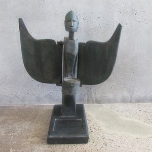 May include: A wooden sculpture of a figure with wings, a head with a carved face, and a small bowl in its hands. The sculpture is painted black and has a simple, minimalist design.