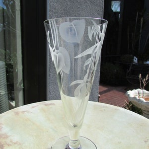 Puede incluir: Un jarr&oacute;n de cristal transparente con la parte superior acampanada y un tallo delgado. El jarr&oacute;n est&aacute; grabado con un dise&ntilde;o floral con hojas y flores. El jarr&oacute;n est&aacute; sobre una mesa redonda de color claro.