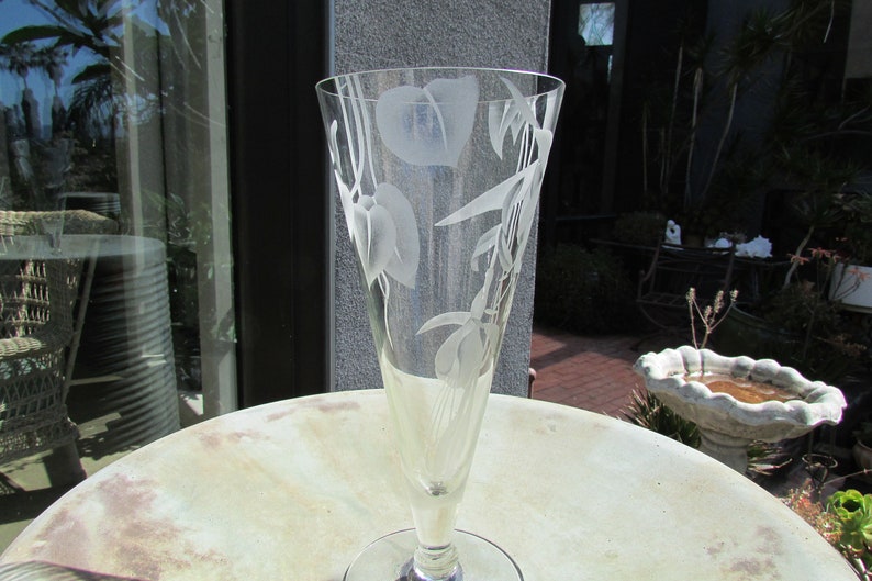 Puede incluir: Un jarr&oacute;n de cristal transparente con la parte superior acampanada y un tallo delgado. El jarr&oacute;n est&aacute; grabado con un dise&ntilde;o floral con hojas y flores. El jarr&oacute;n est&aacute; sobre una mesa redonda de color claro.