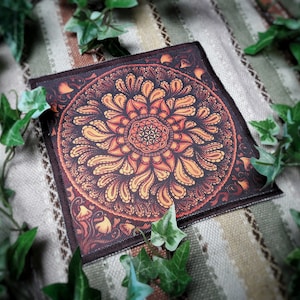 May include: Square, dark brown fabric with a detailed, symmetrical mandala design in shades of orange and yellow. The mandala features a central flower-like pattern and intricate, swirling details. The fabric is surrounded by green ivy.