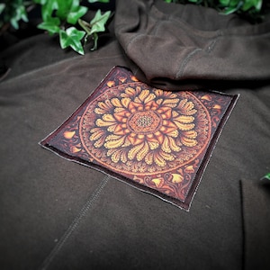 May include: A brown hooded sweatshirt with a square patch featuring a detailed orange and yellow mandala design.