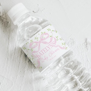 May include: A clear plastic water bottle with a white cap and a label that reads "Katelyn's Baby Shower" in pink script. The label features a pink bow and floral design. A matching label lies below.