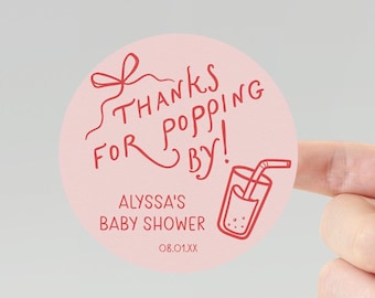 May include: A circular, pale pink sticker featuring red text and a drink graphic. The text says "Thanks for popping by!" and "Alyssa's Baby Shower" with a date. The sticker is held by a finger.