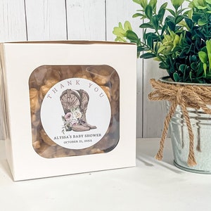 May include: White favor box with a clear window showing brown and tan snacks. A round sticker with the text "Thank You" and a pair of brown cowboy boots with pink flowers is on the front of the box. The text "Alyssa's Baby Shower October 21, 20XX" is on the sticker.