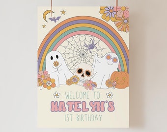 May include: A vintage-style birthday party sign featuring a rainbow, ghosts, a skull, and a jack-o'-lantern. The sign reads "Welcome to Katelyn's 1st Birthday" in pink and purple text. Flowers and a spiderweb complete the design.
