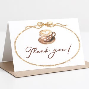 May include: A folded white card with a watercolor illustration of a coffee cup with a heart-shaped design, inside a gold oval frame. The words "Thank you!" are written in brown script below the frame. The card is on a light brown envelope.