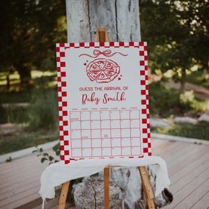 May include: A white sign with a red checkered border and a pizza graphic at the top. The text "GUESS THE ARRIVAL OF Baby Smith" is displayed above a calendar grid. The sign is mounted on a wooden easel.