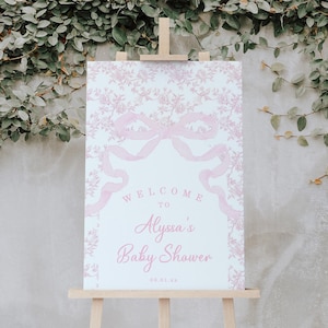 May include: A pink and white welcome sign with a floral pattern and a large pink bow. The text reads "Welcome to Alyssa's Baby Shower 08.01.xx".