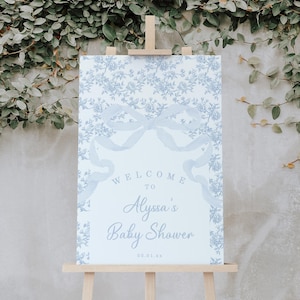 May include: A baby shower welcome sign on a wooden easel. The sign features a light blue floral pattern and a large bow design. Text reads "Welcome to Alyssa's Baby Shower" with a date below. The background is a concrete wall with greenery.