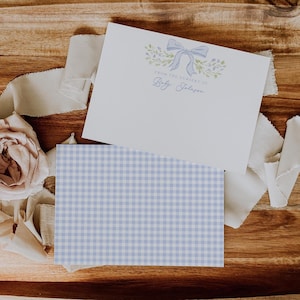 May include: Two stationery cards. The top card is white with a blue bow and floral design, and the text "FROM THE NURSERY OF Baby Johnson". The bottom card has a blue and white gingham pattern. Ribbons and a rose are in the background.