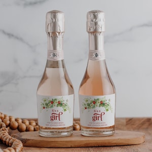 May include: Two mini bottles of sparkling wine with labels that say "It's a girl" and "Pop to open when baby Johnson arrives". The labels feature a floral design with strawberries.