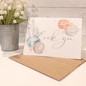 May include: A white thank you card with a watercolor illustration of planets with faces. The planets are in shades of blue, green, orange, and gray. The card has the words "Thank you" written in black cursive.