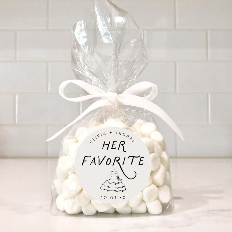May include: A clear cellophane bag filled with white marshmallows, tied with a white ribbon. A circular sticker on the bag reads "Olivia + Thomas, Her Favorite" with a dress illustration and the date "10.01.XX".