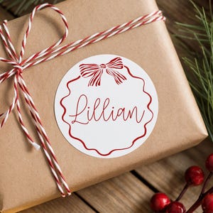 May include: A wrapped gift in brown paper, tied with red and white twine. A white circular sticker with a red bow design and the name "Lillian" in red script is affixed to the top of the package. The background includes greenery and berries.