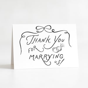 May include: A white folded card with black handwritten text that reads "Thank You for Marrying Us!" The card features a decorative bow and two wedding rings with a diamond.