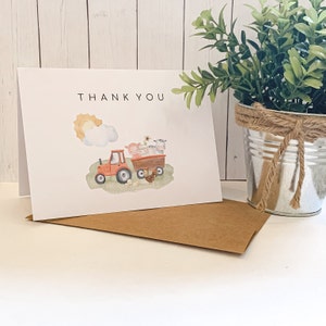 May include: A white thank you card with a red tractor carrying farm animals. The tractor is pulling a wagon with a cow, pig, and sheep inside. The sun is shining in the background.