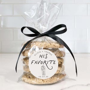 May include: A clear cellophane bag tied with a black satin ribbon, containing several chocolate chip cookies. A white circular sticker on the bag reads "Olivia + Thomas His Favorite" with a tuxedo graphic and the date "10.01.XX".