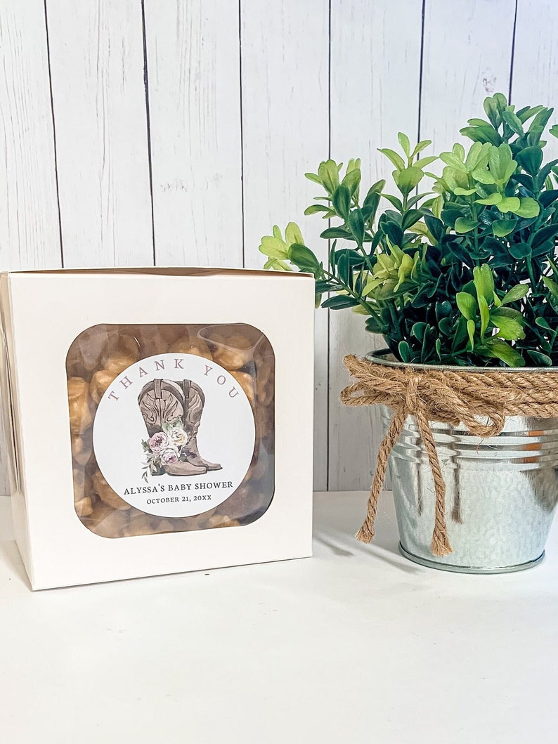 May include: White favor box with a clear window showing brown and tan snacks. A round sticker with the text "Thank You" and a pair of brown cowboy boots with pink flowers is on the front of the box. The text "Alyssa's Baby Shower October 21, 20XX" is on the sticker.