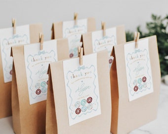 May include: Brown paper bags with thank you tags attached with clothespins. The tags have a blue and pink donut design and say "Thank you for celebrating with us Alyssa and baby donuts".