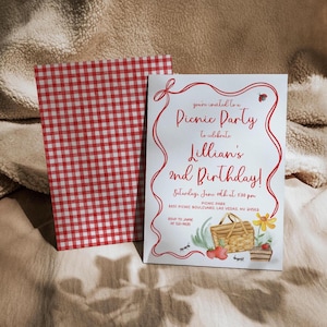 May include: A red and white checkered picnic blanket with a white invitation card on top. The invitation card has red text that says "You're invited to a Picnic Party to celebrate Lillian's 2nd Birthday!" The invitation also includes a date, time, and location for the party. There is a drawing of a picnic basket, strawberries, and ants on the card.