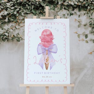 May include: Welcome to Lillian's First Birthday sign with a pink and purple ice cream cone with a bow on top. The sign has a pink and white background with a pink border.