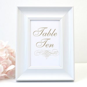 May include: A white framed table number sign with the text "Table Ten" in a gold script font. The sign has a decorative flourish at the bottom. A bouquet of pink flowers is in the lower left corner.