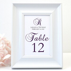 May include: White framed table number sign with purple text. The sign reads "Sarah & Thomas February 14, 2018" and "Table 12". A pink flower arrangement is in the background.