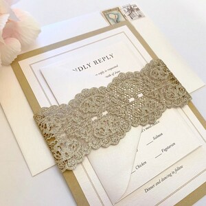 May include: An invitation suite featuring a gold lace band wrapped around a white card with text. The card is inside a kraft paper envelope, with a white envelope behind it. Two postage stamps are visible.