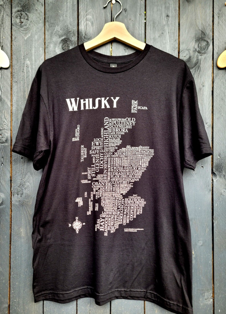 May include: Black t-shirt with a white print of a map of Scotland. The map is made up of words related to whisky, including distillery names, regions, and types of whisky. The word "WHISKY" is printed at the top of the shirt.