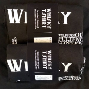 May include: Two black t-shirts with white text. The text on the shirts reads "Whisky T-shirt" and includes the names of different whisky brands: Wolfburn, Pulteney, and Clynelish. The shirts are folded and stacked on top of each other.