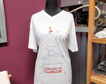 May include: A white V-neck t-shirt featuring a black dot-to-dot graphic of a statue wearing an orange traffic cone on its head. Below the graphic, the text I BELONG TO GLASGOW is printed in red capital letters on the front of the garment.