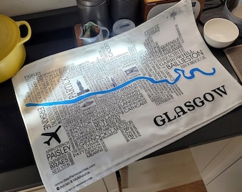 May include: A white tea towel with a blue and black map of Glasgow, Scotland. The map includes the names of neighborhoods and landmarks. The text "GLASGOW" is printed in large black letters at the bottom right corner.