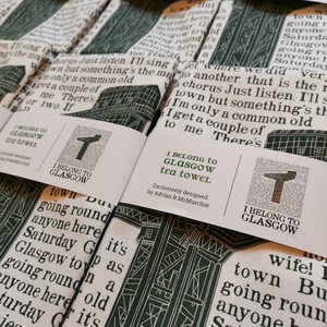 Op de afbeelding: Een stapel witte theedoeken met een groen-wit grafisch ontwerp. De theedoeken bevatten de tekst "I Belong to Glasgow" en een gestileerde letter "T".