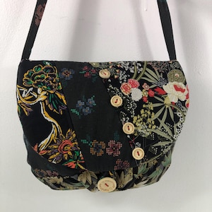 Peut inclure: Un sac bandoulière noir avec un motif floral dans diverses couleurs, dont le jaune, le rouge et le blanc. Le sac est orné de boutons en bois et d'une sangle noire. Le sac est en tissu.