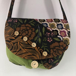 May include: A patchwork crossbody bag with a flap closure. The bag features floral patterns in brown, black, and cream, and a solid green base. It has a long strap and is secured with wooden buttons. The word "handmade" is printed on the buttons.