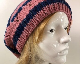 May include: A pink and navy blue knitted beret with a striped pattern. The hat has a textured appearance and a small wooden button detail. The beret is worn on a mannequin head with blonde hair.