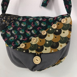 Peut inclure: Sac bandoulière fait main, style patchwork. Il présente une base grise, un rabat avec des motifs floraux et géométriques verts foncés, dorés et noirs, et des boutons en bois. La bandoulière est en tissu à motifs assortis.