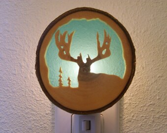 May include: A wooden night light shaped like a deer head with a teal blue background. The deer head is cut out of the wood and the light shines through the cutout.