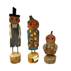May include: Three wooden pumpkin-headed figures. The tallest figure wears a black top hat and overalls. The middle figure has a sunflower on its dress. The smallest figure wears a scarf and a vest. All figures are mounted on wooden bases.