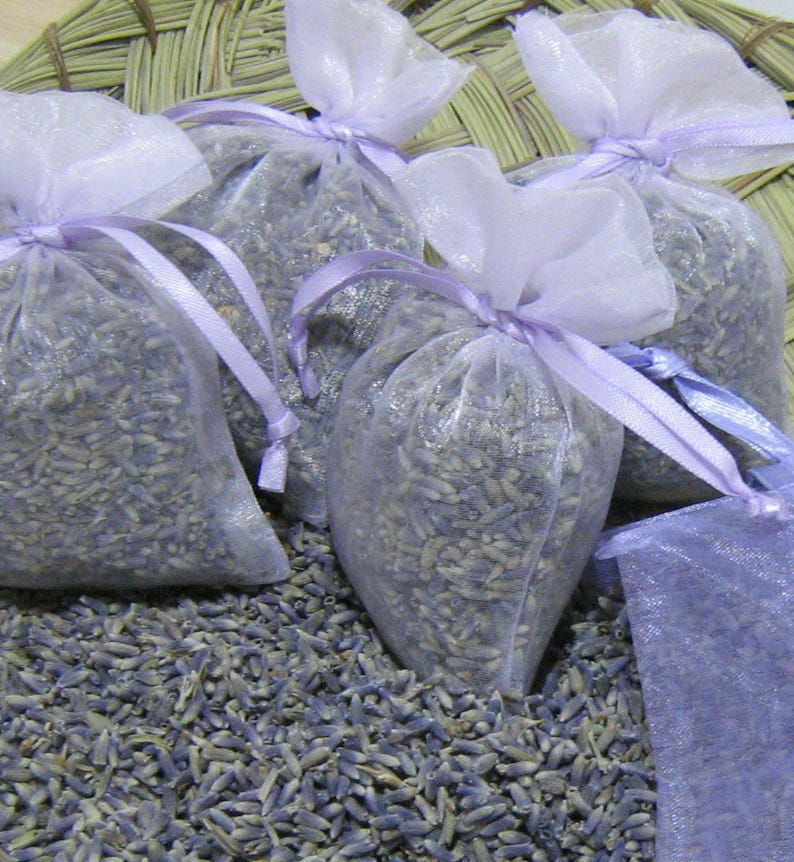 May include: Four lavender-scented sachets made of sheer purple fabric with lavender buds inside. The sachets are tied with purple ribbon and are sitting on a bed of dried lavender.