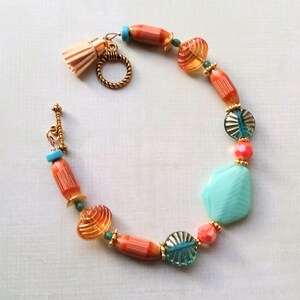 May include: A beaded bracelet featuring a variety of colorful beads. The bracelet includes orange, teal, and gold-colored beads, along with a large turquoise stone. A small tassel and gold-colored clasp complete the design. The bracelet is approximately 7 inches in length.
