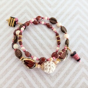 Op de afbeelding: Een meerlagige armband met een mix van bruine, crème en roze kralen. De armband bevat koffieboonvormige kralen, kleine gouden kralen en decoratieve bedels, waaronder een kleine, roze en zwarte bedel. De armband ligt op een wit en grijs chevron patroon.