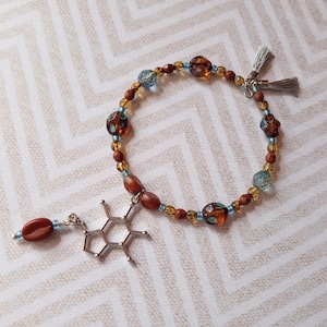 Op de afbeelding: Een kralenarmband met bruine, blauwe en oranje kralen. Een zilveren bedel van een cafeïnemolecuul en een koffieboon hangen aan de armband. De armband heeft een kwastjessluiting.