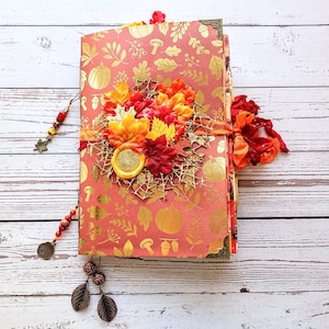 Puede incluir: Un diario hecho a mano con temática otoñal. La portada está decorada con un grupo de hojas naranjas, rojas y amarillas, una calabaza dorada y un patrón de hojas doradas. El diario está encuadernado con una cinta marrón y tiene un borla de cuentas.