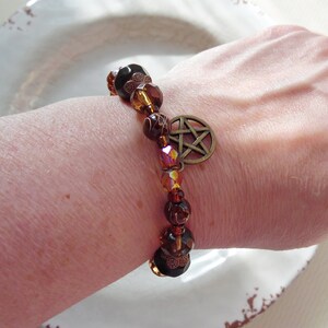 May include: A beaded bracelet featuring a bronze pentagram charm and a mix of brown, amber, and iridescent beads. The bracelet is made with various sizes and shapes of beads, creating a layered effect. The bracelet is worn on a wrist.