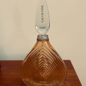May include: A heart-shaped amber glass perfume bottle with a clear teardrop-shaped stopper featuring the text CHAMADE. The bottle has a ribbed texture, a thin cord wrapped around the neck, and a base with the text NET CONT 4 FL OZ printed on it.
