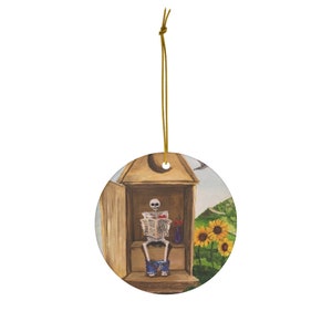 May include: A round ceramic ornament featuring a cartoon skeleton sitting on a toilet reading a newspaper. The toilet is inside a wooden outhouse with a sunflower and a green hill in the background.